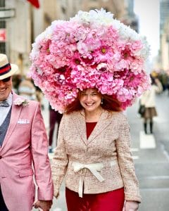 A woman smiling while wearing an extravagant Easter bonnet covered in pink and white flowers at the Easter Bonnet Parade in New York City. She walks hand-in-hand with a man dressed in festive attire, surrounded by the lively atmosphere of the annual Easter Sunday celebration.