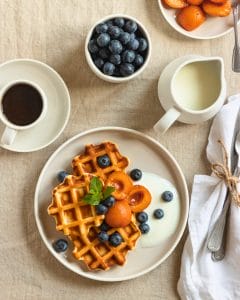 A plate of golden waffles topped with fresh blueberries, caramelized apricots, and a dollop of yogurt, garnished with a sprig of mint. Surrounding the plate are a cup of coffee, a jug of milk, a bowl of blueberries, and a dish of apricots, all set on a neutral-toned linen tablecloth.