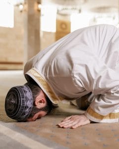 A man in traditional white and gold attire performing sujood (prostration) during prayer inside a mosque, symbolizing the Eid al-Fitr prayer.