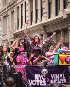 A lively group of people in colorful outfits and accessories dance and celebrate on a parade float decorated with “NYC Votes x Vinyl Nights” signage. One person dressed as the Statue of Liberty holds a torch aloft, surrounded by music-themed props and confetti, with buildings lining the background.