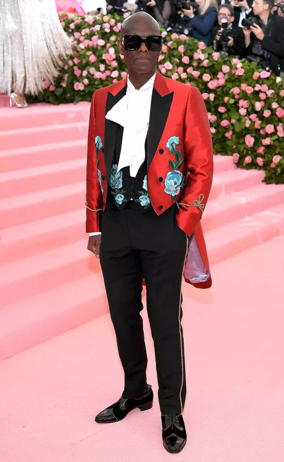 Dapper Dan poses at the Met Gala in a red embroidered tuxedo jacket with blue floral detailing, black trousers, and oversized sunglasses. Image from People.com for the blog post "Met Gala 2025: Celebrities, Red Carpet, Theme & More."