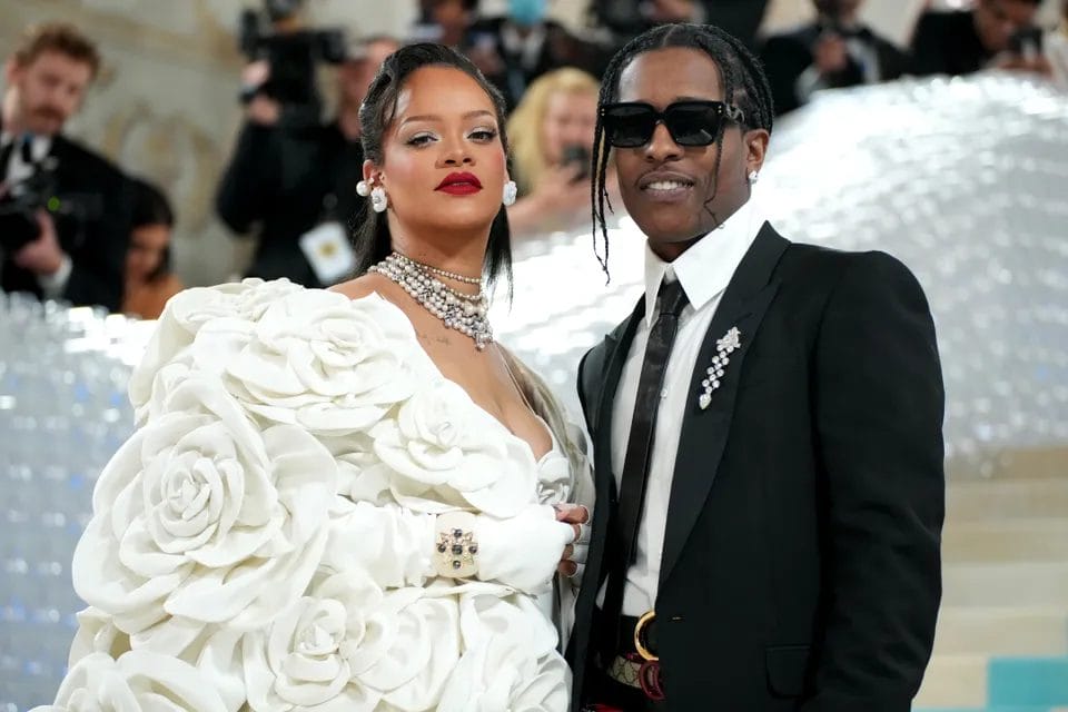Rihanna and A$AP Rocky pose at the Met Gala; Rihanna wears a dramatic white gown adorned with large floral rosettes, while A$AP Rocky wears a classic black suit with statement pins. Featured in the blog post "Met Gala 2025: Celebrities, Red Carpet, Theme & More."