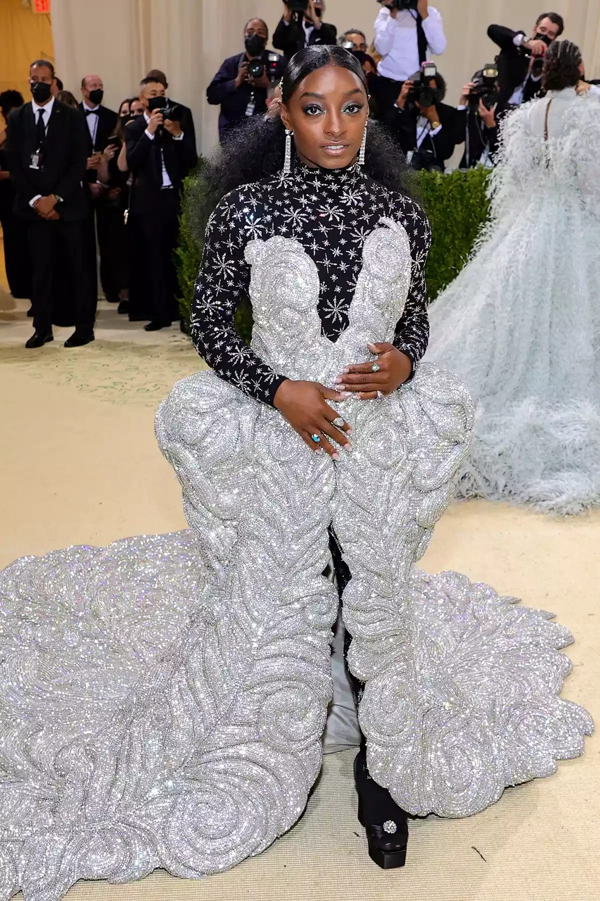 Simone Biles poses on the Met Gala steps in a black and silver embellished gown with a dramatic, sculpted silver train. Featured in the blog post "Met Gala 2025: Celebrities, Red Carpet, Theme & More."