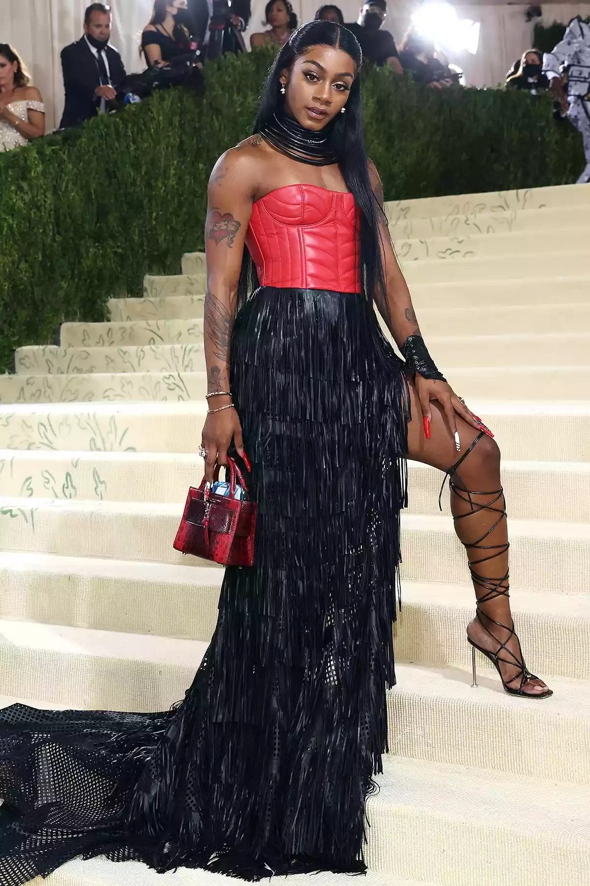 Sha'Carri Richardson poses at the Met Gala in a bold look featuring a red leather corset, black fringe skirt, lace-up heels, and a red handbag. Featured in the blog post "Met Gala 2025: Celebrities, Red Carpet, Theme & More."