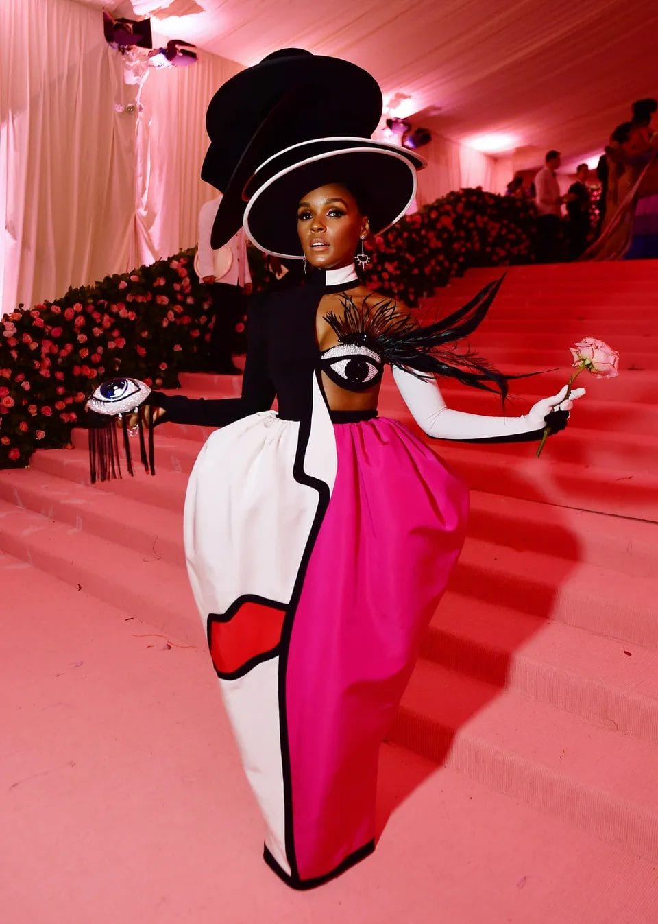 Janelle Monáe at the Met Gala in a bold black, white, and pink sculptural gown featuring abstract facial art, a feathered eye design, and stacked wide-brim hats.