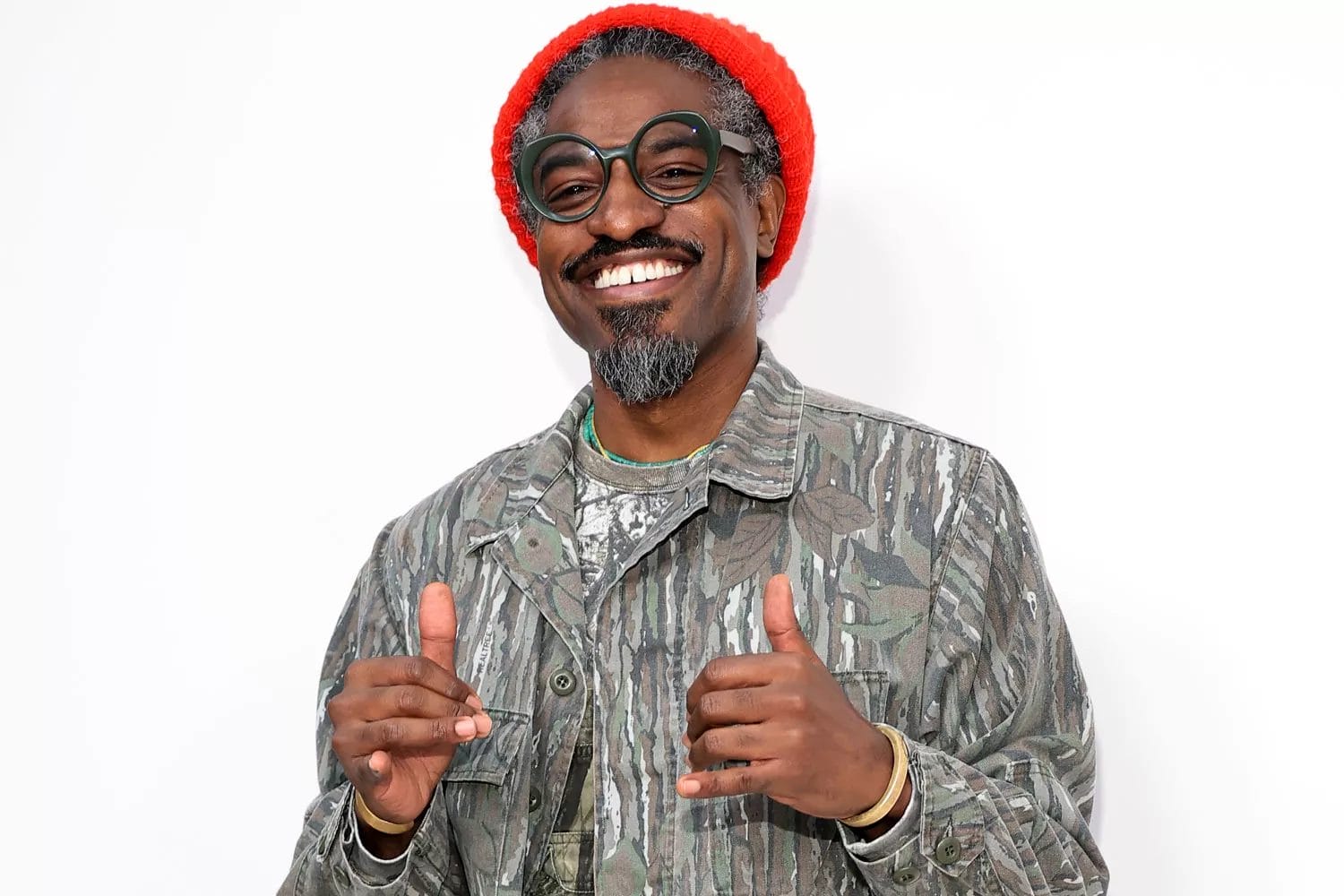 André 3000 smiles in a camouflage jacket and red beanie, wearing oversized glasses and posing with both thumbs up.
