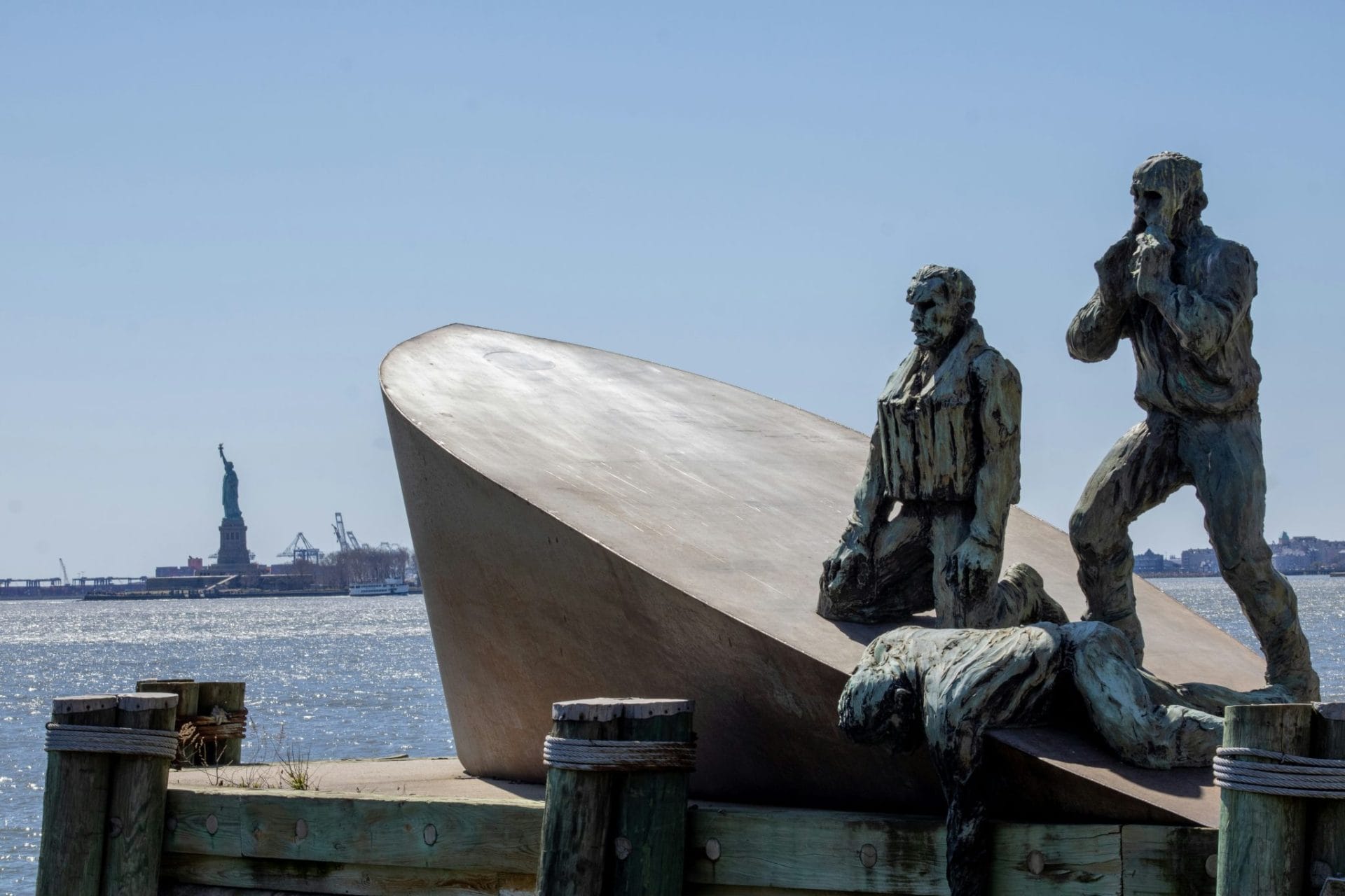 Wide shot of edge of Battery Park, with a statue in focus with the Statue of Liberty in the back.