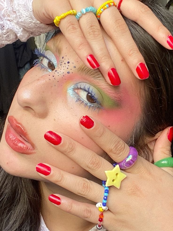 A person with colorful makeup, red painted nails, funky and colorful rings.
