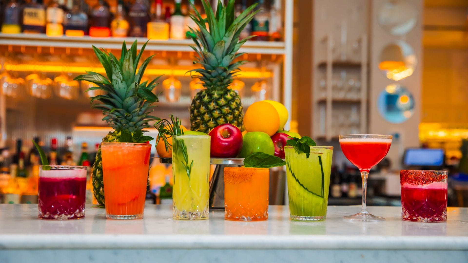A vivid row of diverse colorful cocktails set on a marble bar top, flanked by two whole pineapples. The drinks include pink, orange, yellow, and green beverages in various glassware, garnished with mint sprigs, rosemary, and sliced cucumber. Behind the cocktails, a silver pedestal stand holds fresh whole fruits including red and green apples, citrus fruits, and oranges. The warm, softly lit bar shelves stocked with bottles create a blurred background.