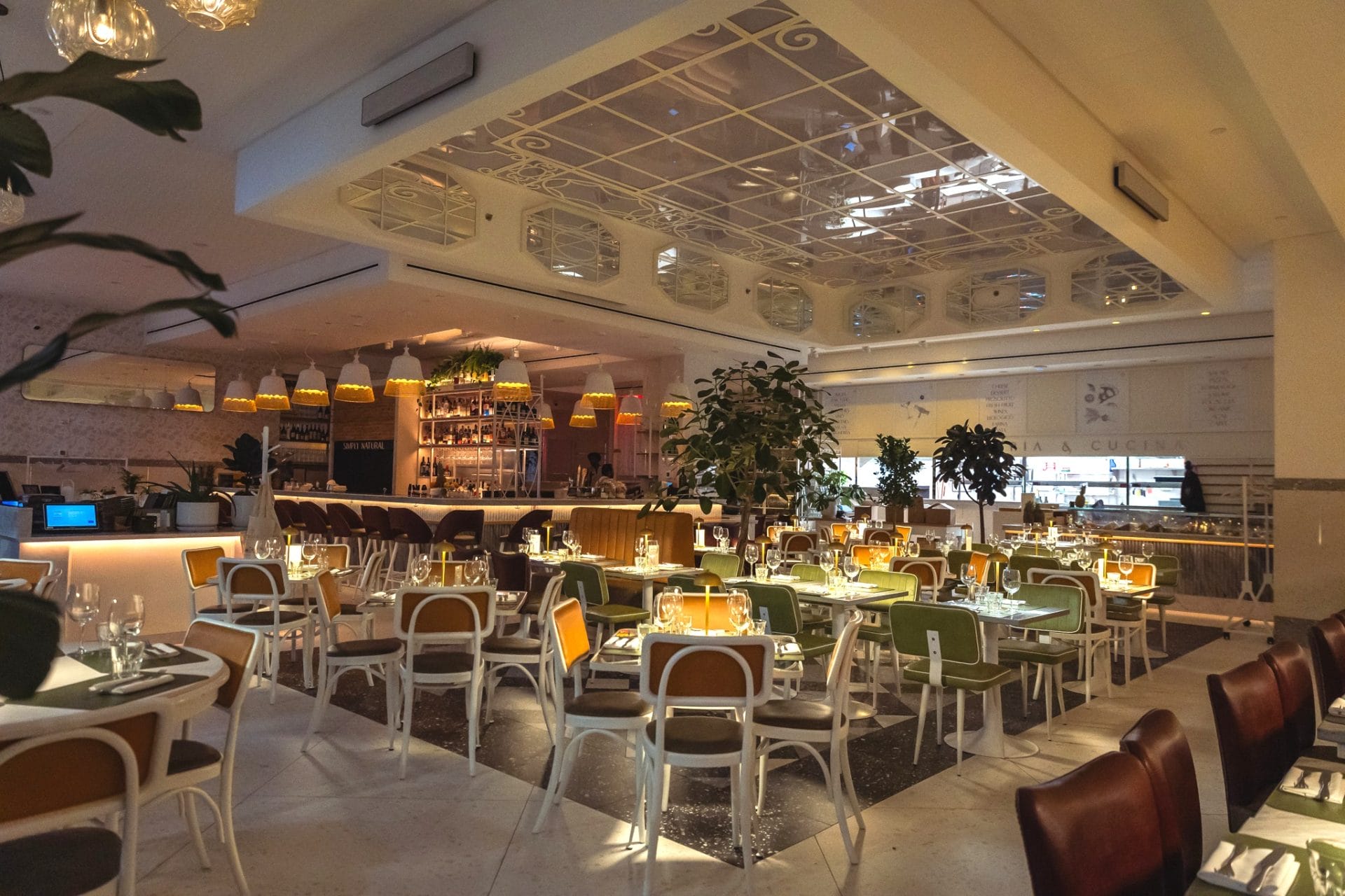 A wide-angle photo showing the main dining room of a modern, spacious restaurant. The space is filled with neatly arranged dining tables paired with white-framed chairs featuring brown or green upholstery. Above the central seating area, an ornate coffered ceiling element with glass panels and decorative scrollwork reflects the warm interior lighting. In the background, a bar counter is illuminated by a row of yellow pendant lights, and indoor leafy potted trees are scattered between table sections.