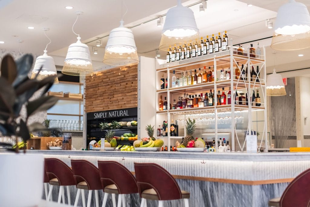 A close-up view of a bar counter featuring a row of sleek, dark maroon barstools lining a light marble top. Hanging above are large white dome pendant light fixtures, and behind the bar stands a multi-tiered white shelving display filled with liquor bottles and fresh fruit displays.