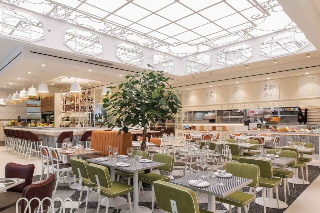 A wide view of a modern restaurant dining room featuring olive-green upholstered chairs, white tables, and potted indoor trees. The space is illuminated by a large ceiling fixture with white glass panels and scrollwork patterns, with a bar and kitchen counter visible in the background.