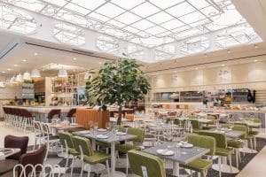 A wide view of a modern restaurant dining room featuring olive-green upholstered chairs, white tables, and potted indoor trees. The space is illuminated by a large ceiling fixture with white glass panels and scrollwork patterns, with a bar and kitchen counter visible in the background.