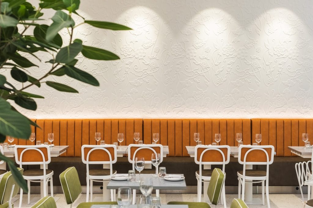 A clean view of restaurant dining tables set with wine glasses and cutlery in front of a long, warm orange leather banquette. White-framed modern chairs surround the tables, positioned against a white feature wall with intricate floral relief patterns illuminated by overhead spotlights.
