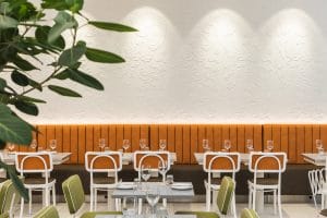 A clean view of restaurant dining tables set with wine glasses and cutlery in front of a long, warm orange leather banquette. White-framed modern chairs surround the tables, positioned against a white feature wall with intricate floral relief patterns illuminated by overhead spotlights.