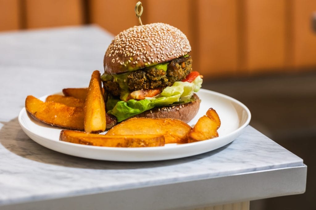 A veggie burger held together with a wooden skewer inside a toasted sesame seed bun, layered with lettuce, tomato, and green pesto sauce. It is served on a white plate alongside golden-brown, thick-cut potato wedges on a marble table.