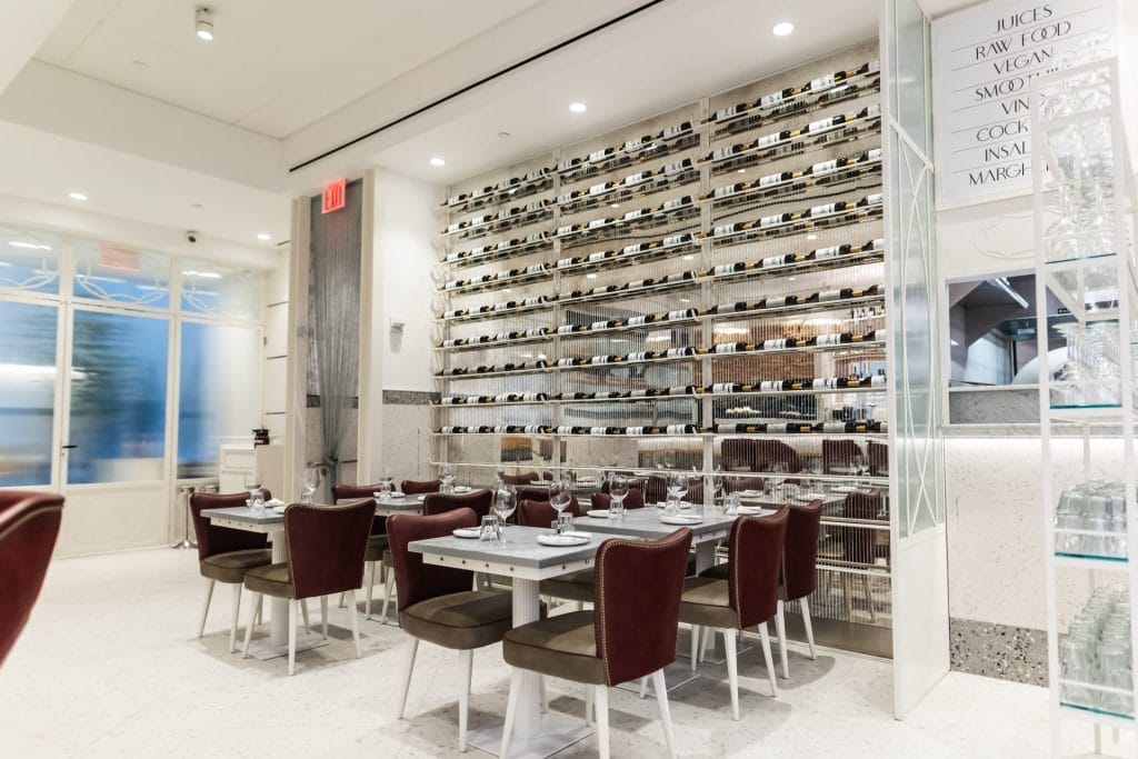A bright restaurant seating area with white marble flooring, set with square tables and plush maroon upholstered chairs. In the background, a large metal rack displays horizontal rows of wine bottles against a mirrored wall.