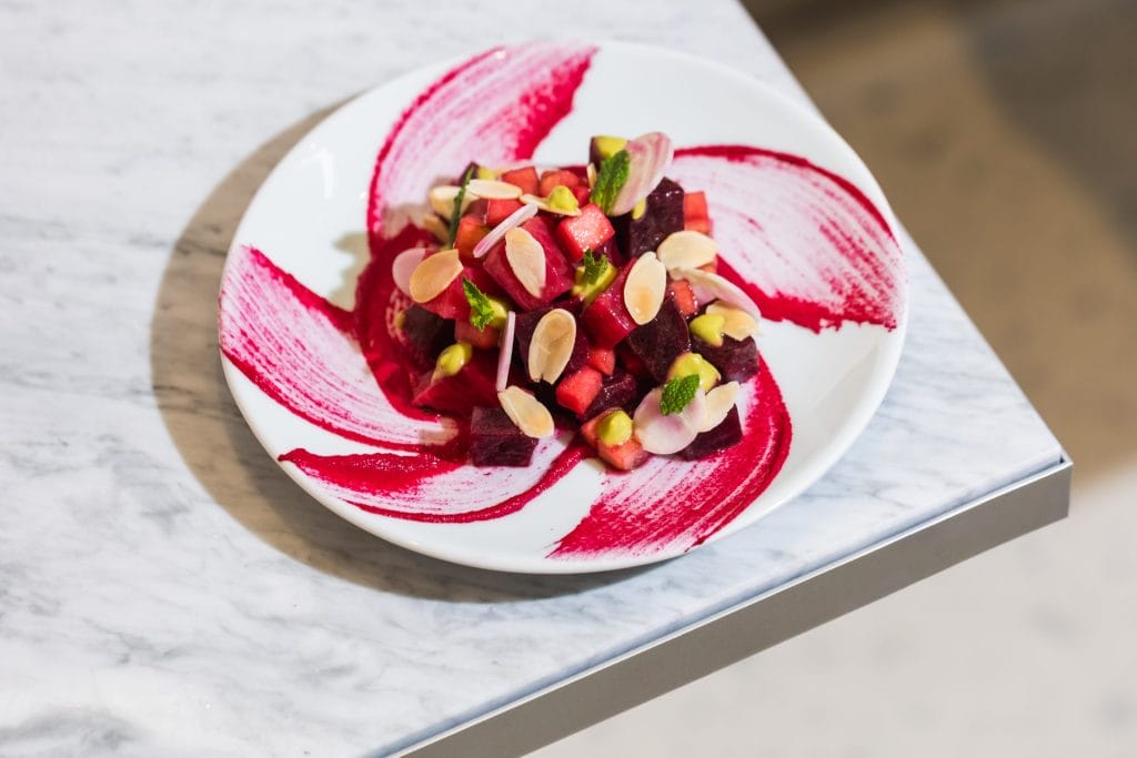 A white plate featuring a decorative swirl of vivid magenta beetroot sauce. At the center is a mound of diced red beets, crisp apple cubes, green pistachios, mint leaves, and flaked almonds.