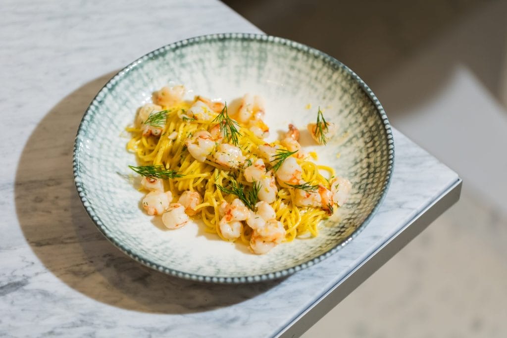 A top-down view of a shallow green pattern bowl containing long, thin yellow pasta mixed with small pieces of sautéed shrimp and garnished with fresh dill sprigs, resting on a white marble tabletop.