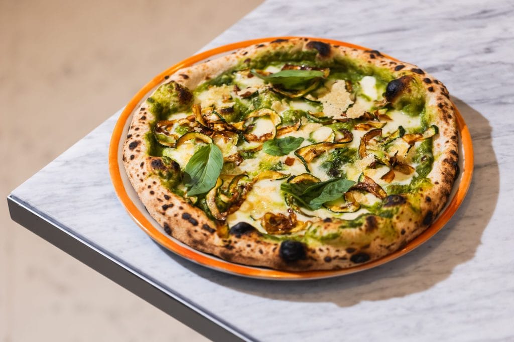 A whole Neapolitan pizza served on an orange-rimmed plate over a light marble surface. The pizza has a charred, puffed crust, a vibrant green pesto base, melted white cheese, roasted zucchini slices, and fresh basil leaves.