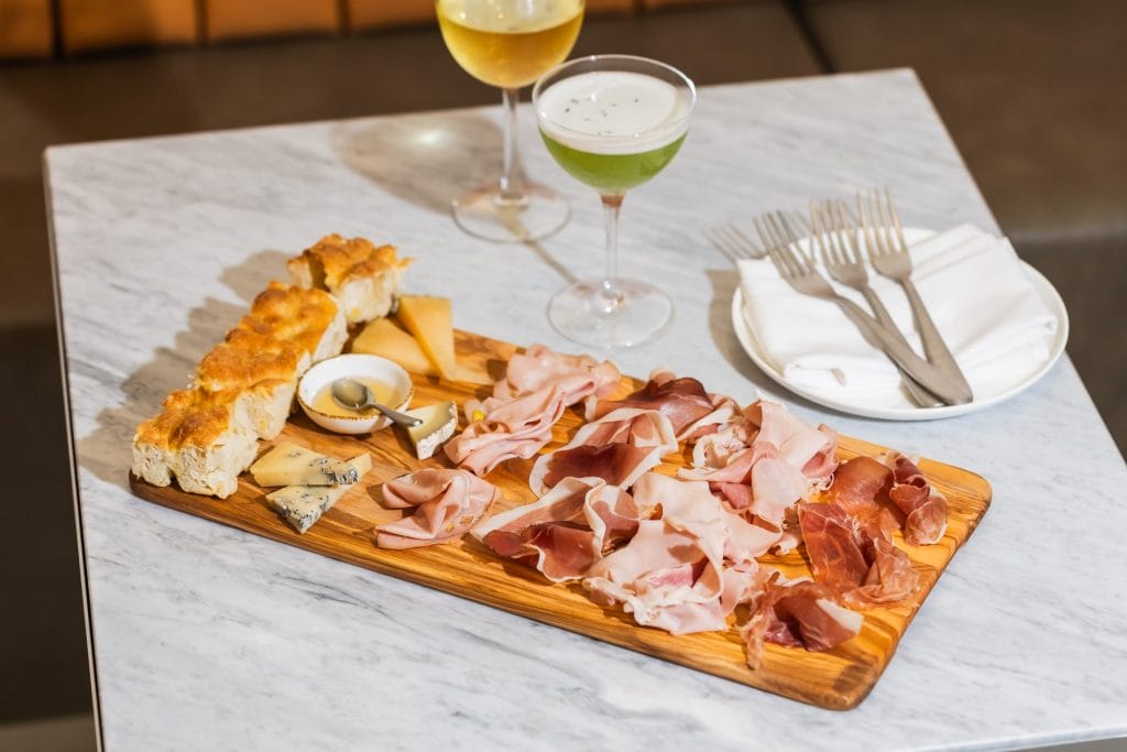 A rectangular wooden board loaded with sliced cured meats, assorted cheese wedges, bread pieces, and a small bowl of honey. Behind the board on the marble table sit two cocktail glasses, three forks, and a folded cloth napkin.