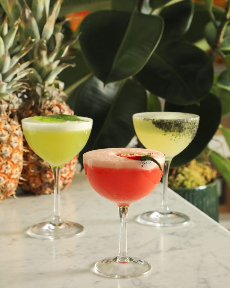 Three distinct cocktails served in stemware coupe glasses arranged on a white marble surface. In the center foreground sits a bright red cocktail garnished with a fresh chili pepper slices. To the left is a light green cocktail topped with white foam and fresh basil leaves, and to the right is a pale yellow cocktail with a dark rim coating. Whole pineapples and lush, dark green broad-leaf houseplants form the background.