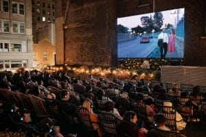 A wide view of an outdoor rooftop cinema venue filled with rows of seated guests wearing headphones, facing a large movie screen displaying a scene from La La Land. Warm decorative string lights line the plant borders below the screen against surrounding city buildings.