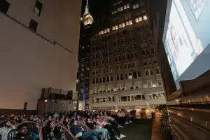 An outdoor rooftop movie screening at night surrounded by tall city buildings, with the illuminated Empire State Building visible in the background sky. Viewers relax in reclined chairs with glowing headphones, facing a large projection screen displaying a film.